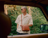 A person dressed in a light summer outfit with a metro crossbody in mahogany, standing amidst lush greenery, captured from the view of a vintage car window, conveying a laid-back, outdoorsy feel.
