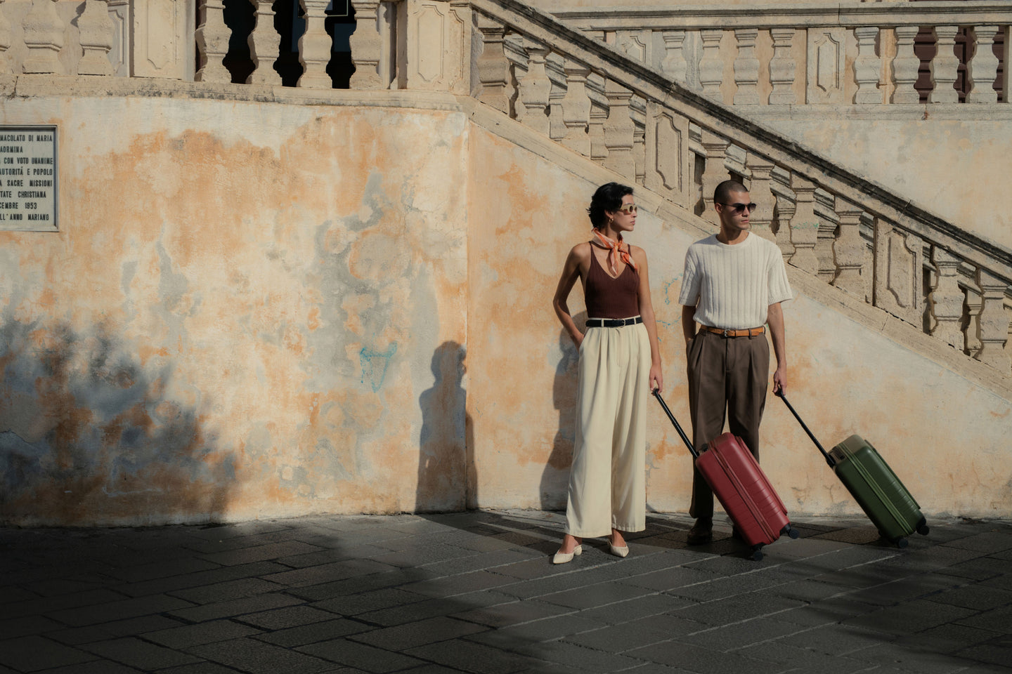A stylish couple standing against a weathered, sunlit wall with a staircase in the background. The woman is wearing a brown sleeveless top and beige wide-leg trousers, holding a carry-on pro in terracotta, while the man wears a white knit top and brown trousers, holding a carry-on pro in olive green. Both are looking off to the side, creating a relaxed yet fashionable travel vibe.
