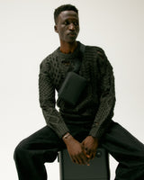 Model sitting on a carry-on suitcase, wearing a Metro Belt Bag in carbon black (vegan leather) across the chest.