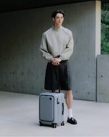A man standing with an expandable carry-on pro in storm grey on a clean concrete surface.