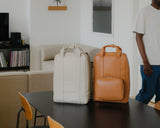 Two vegan leather Metro Backpacks placed on a table. The Metro Backpack in Saddle Tan (Vegan Leather) on the right has the Metro Classic Kit attached to the front, while the Metro Backpack in Ivory (Vegan Leather) on the left does not have the Metro Classic Kit attached.