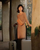 A woman standing in a softly lit corridor next to Monos suitcases in desert taupe. She is wearing a beige-toned dress and a metro sling in saddle tan (vegan leather), gazing off into the distance.