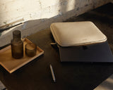 A metro laptop sleeve in ivory (vegan leather) is partially open, revealing a laptop. The laptop sleeve is placed on a dark surface next to a wooden tray with two ribbed glass cups and a silver pen, all lit by soft natural sunlight.