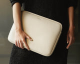 A close-up of a woman's hand holding a metro laptop sleeve in ivory (vegan leather) against a backdrop of a dark sweater.