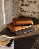 Two metro laptop sleeves in saddle tan (vegan leather), stacked on a dark surface, with one holding a laptop that is slightly visible. The scene is lit by warm natural light, creating a cozy, rustic atmosphere with exposed brick walls in the background.