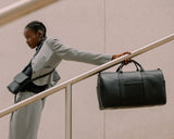 A person dressed in a sleek grey suit leaning against a railing while carrying a Metro Carry-All Duffel in Carbon Black (Vegan Leather) in one hand and a Metro Crossbody Bag in Carbon Black (Vegan Leather). The person is standing on stairs in an urban, minimalist setting.