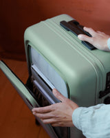 A close-up of hands opening the front pocket of a carry-on in sage green, revealing its organized interior.