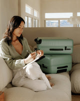 A woman sitting on a couch, writing in a journal, with two stacked check-in suitcases in sage green nearby.
